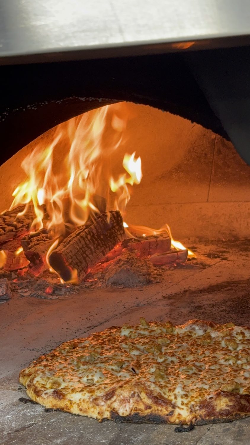 A pizza baking beside the flames in the wood-fired oven